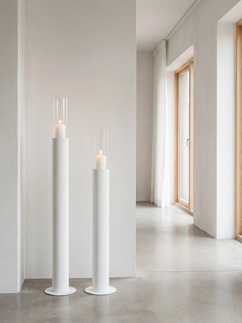 Thomas candleholders in white with glass cylinders and lit candles, placed in a minimalist Scandinavian hallway with concrete flooring, soft white walls, and natural light from wooden-framed windows.