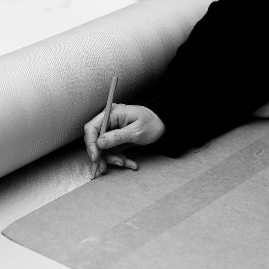 Black-and-white close-up of a craftsman's hand marking upholstery fabric beside a patterned textile roll, capturing the precision and care of handmade furniture production in Sweden.