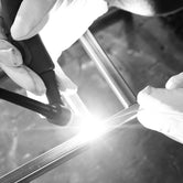 Close-up, black and white shot of hands in protective gloves performing TIG welding on metal pipes, with a bright arc illuminating the joint. Ideal for industrial manufacturing, fabrication, and precision welding content.