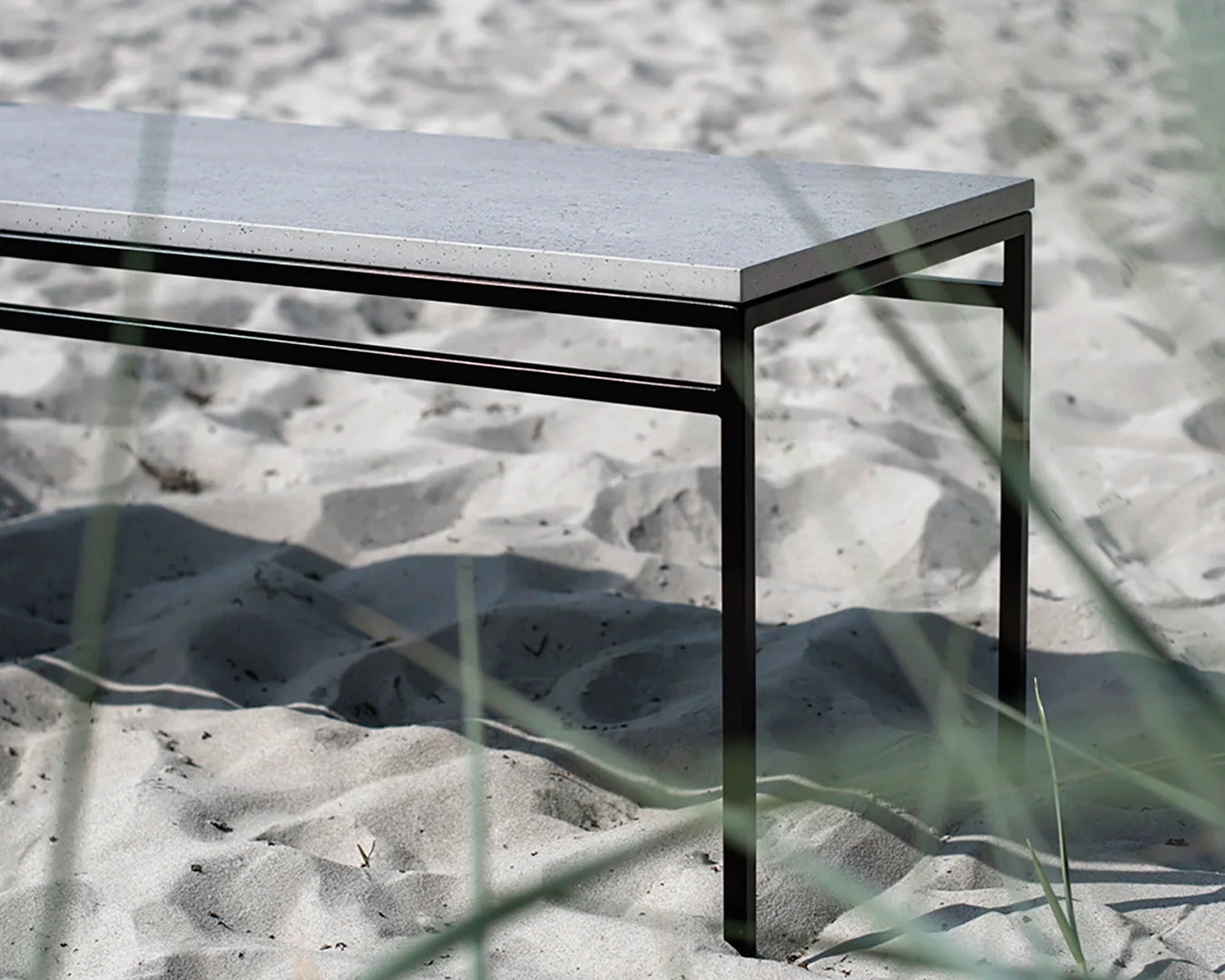 Wilma outdoor bench with a black powder-coated steel frame and durable Dekton stone top, styled in a coastal sand setting with natural grass in the foreground.