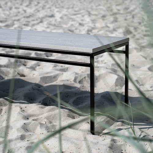 Wilma outdoor bench with a black steel frame and grey Dekton top, placed on a sandy beach with natural dune grass in the foreground.