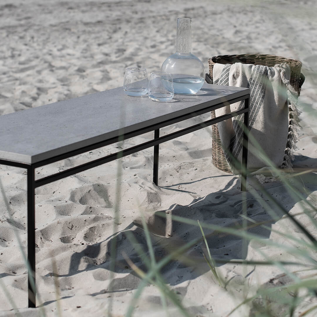 Wilma outdoor bench with sleek Keon dekton top on the beach, complemented by glassware and a woven basket.